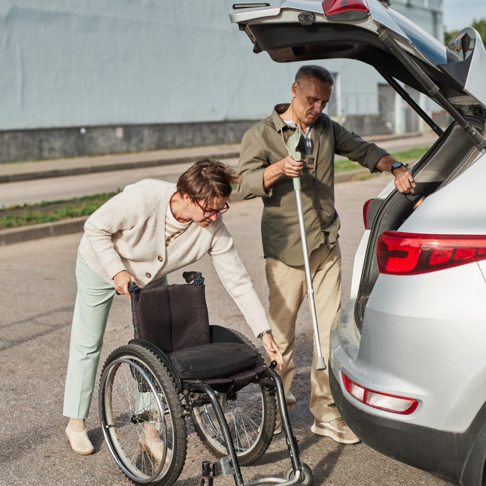 Full length portrait of mature couple loading wheelchair to car trunk in parking lot outdoors