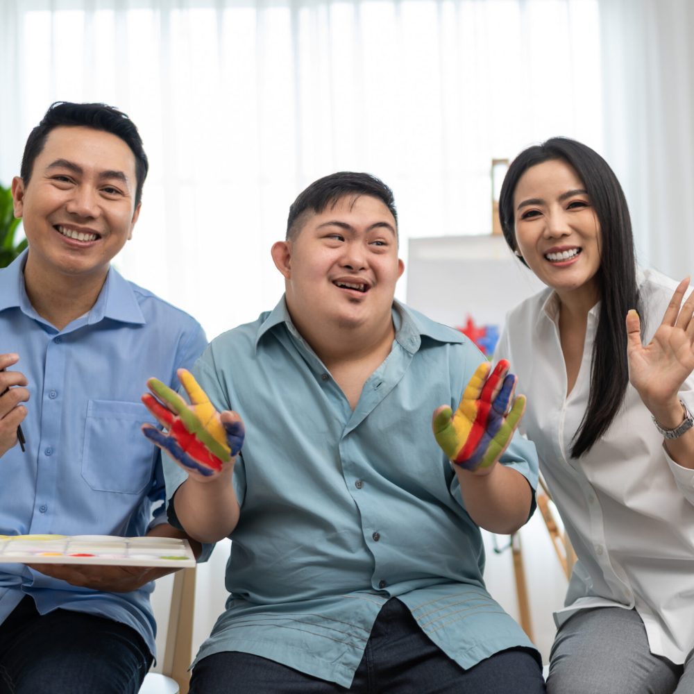Asian happy family draw picture with down syndrome son in living room. Beautiful loving mother and father take care and teach young man with autism or special needs painting artwork indoors at home.