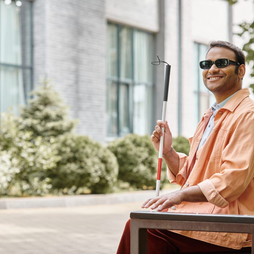 joyful indian blind man in orange jacket sitting outside on bench with walking stick and glasses